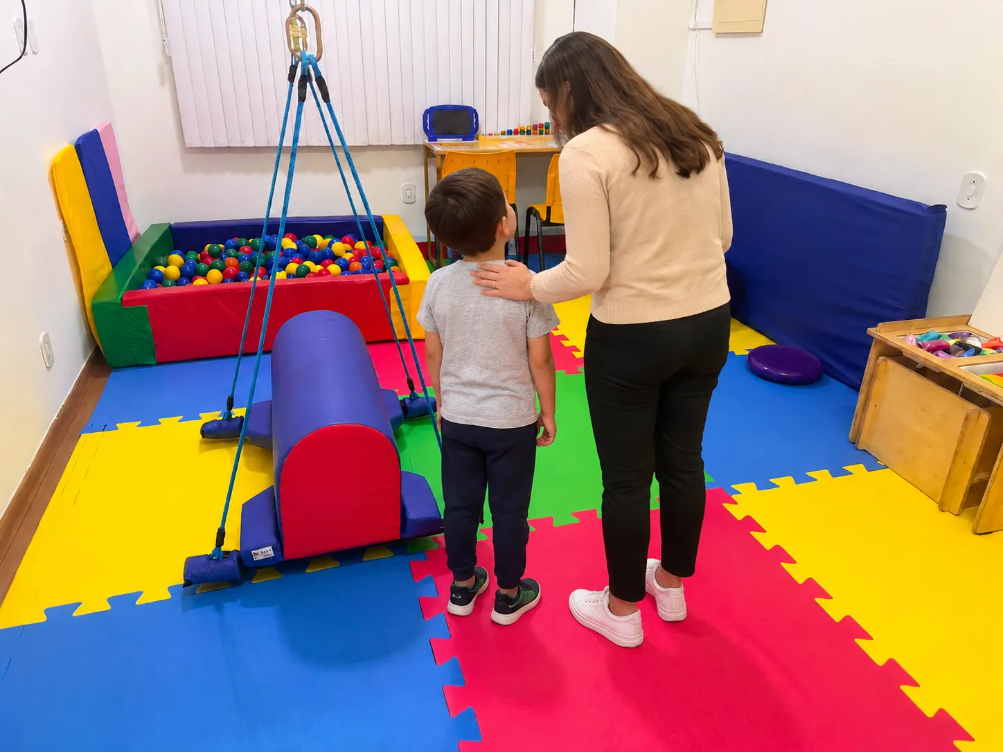 Psicóloga com a mão no ombro de uma criança, ambas de costas, na sala de integração sensorial da Semear com piscina de bolinhas e balanço